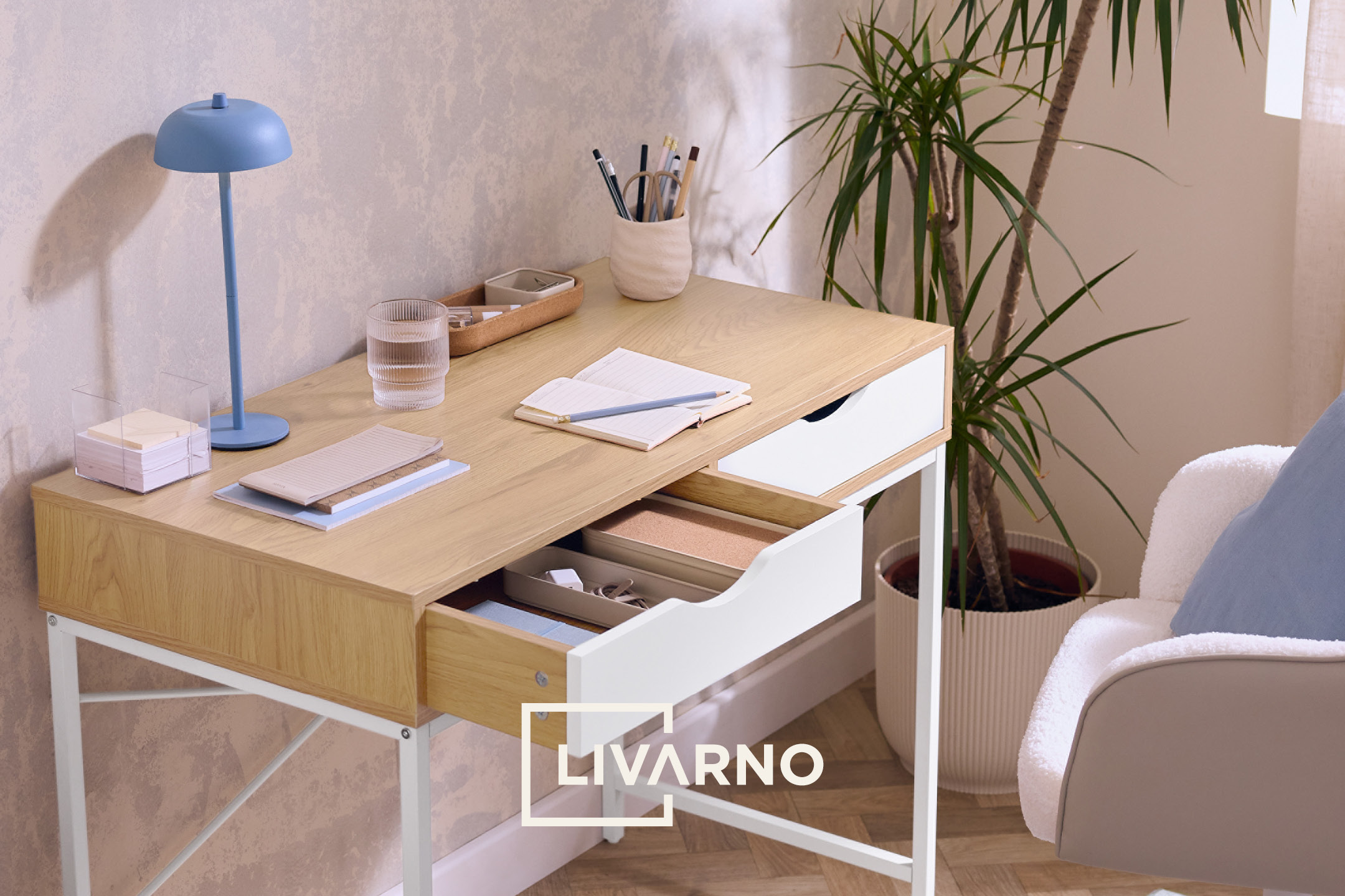 Scrivania in legno chiaro con lampada blu, bicchiere d'acqua, quaderno e portapenne.