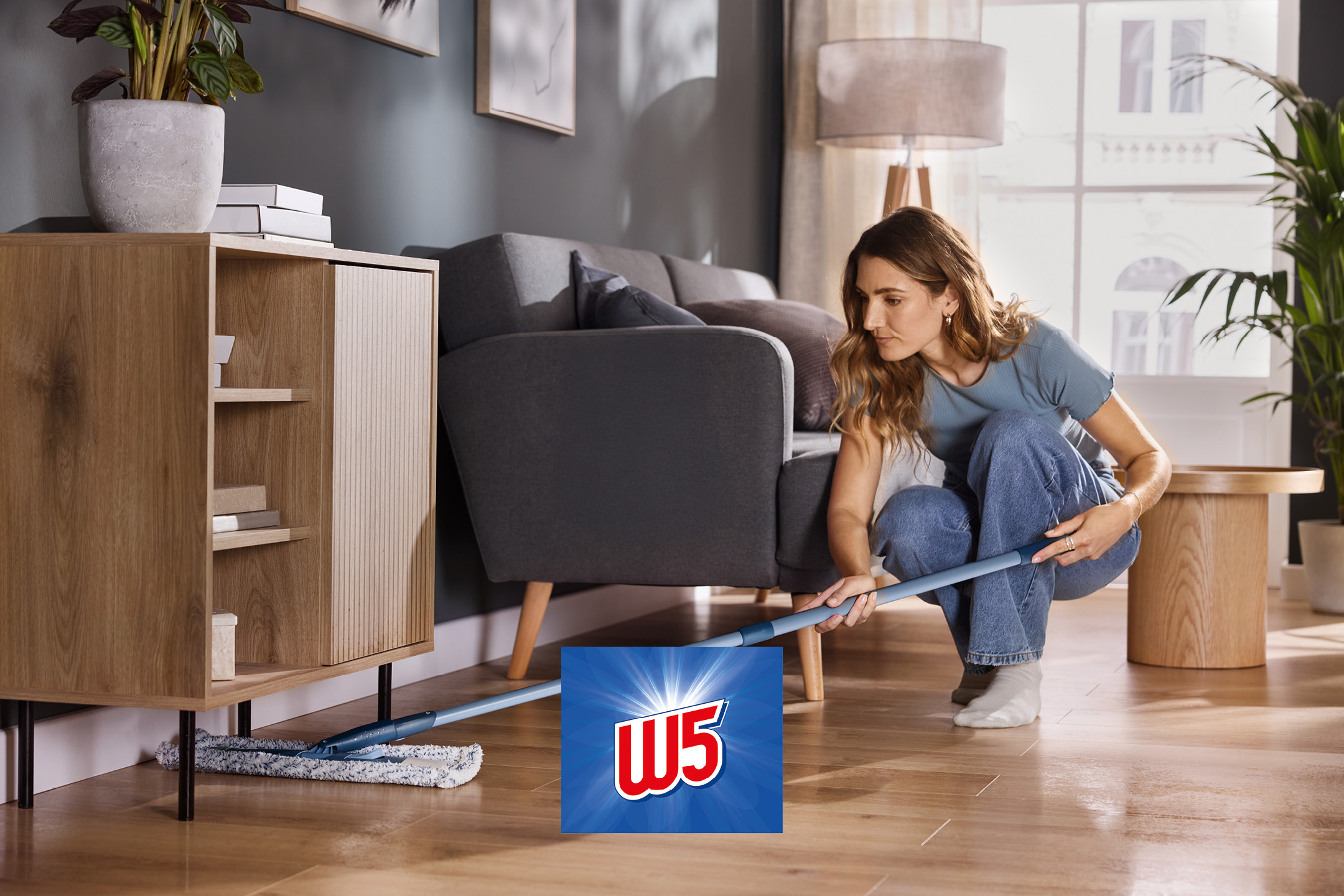 Una persona pulisce il pavimento in legno sotto un mobile con un mocio piatto.