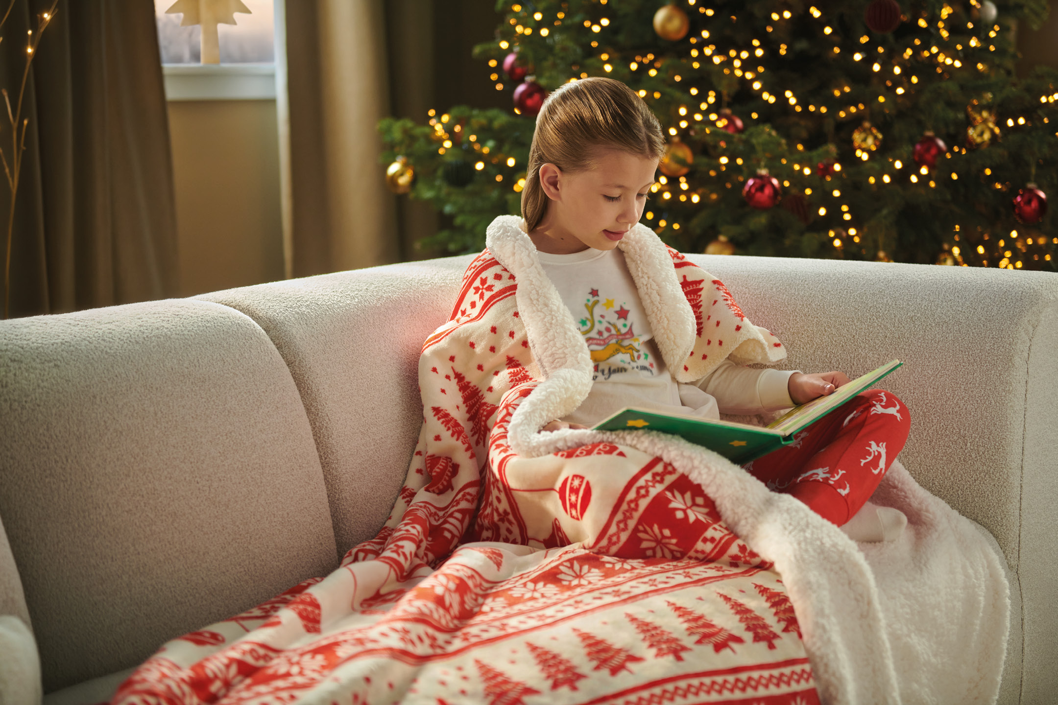 Bambina seduta sul divano con coperta natalizia mentre legge un libro.