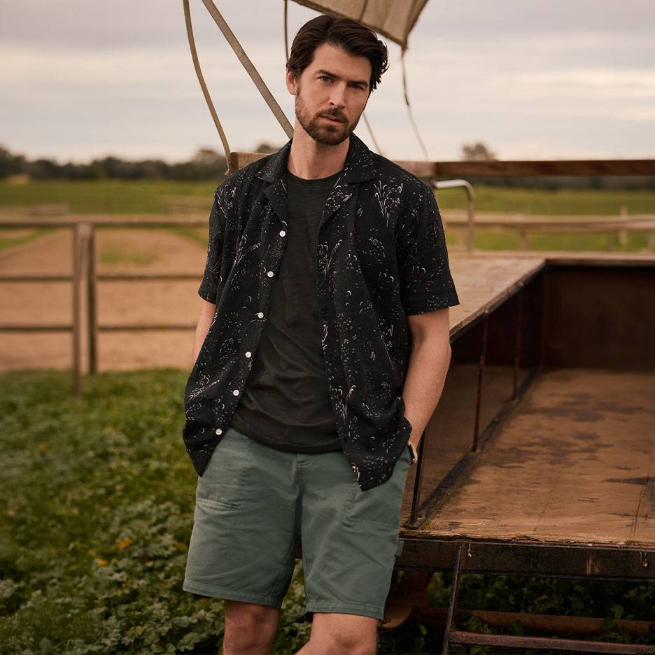 Uomo in camicia nera fantasia, maglietta e pantaloncini verdi, in piedi accanto a un rimorchio.