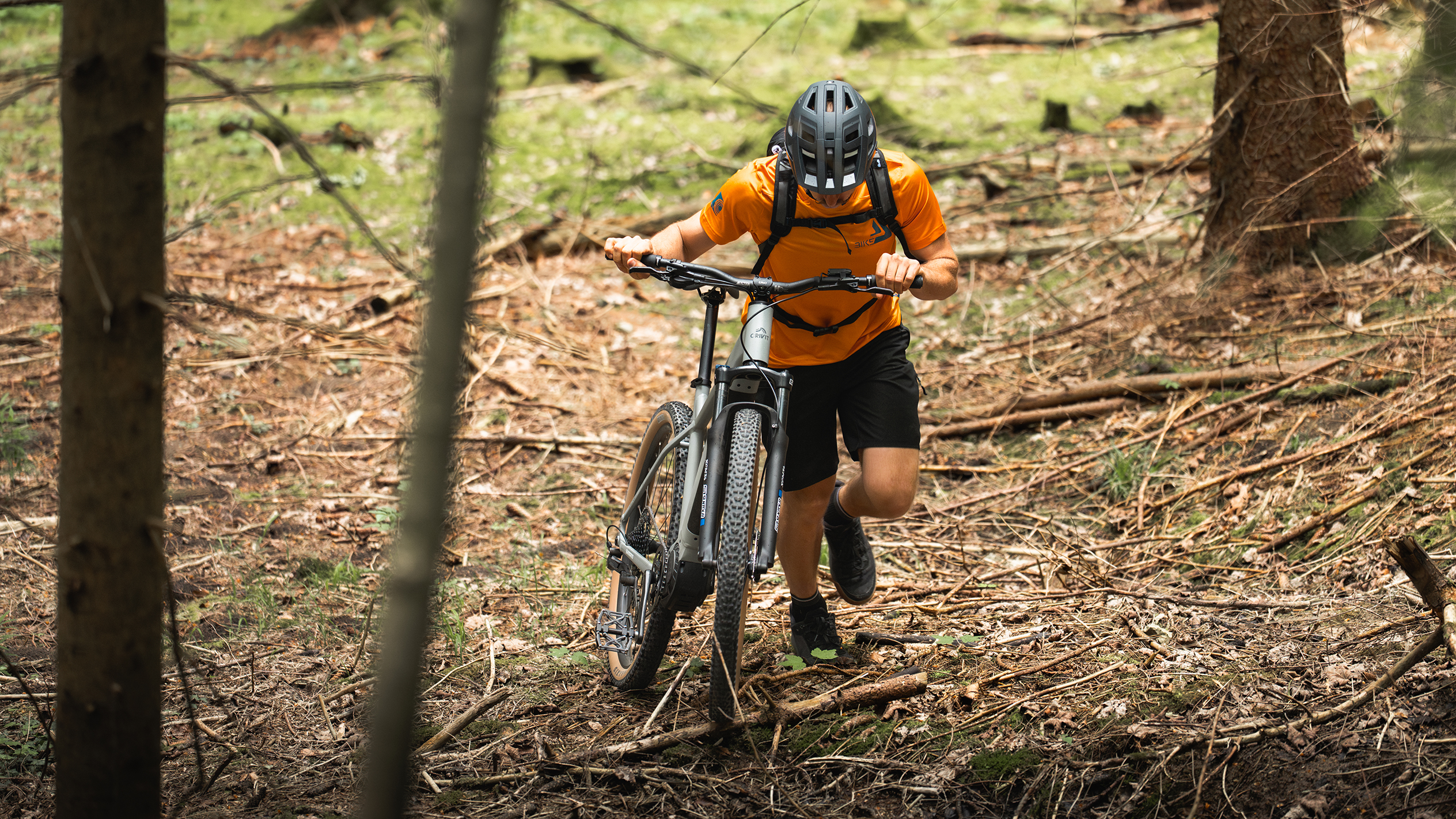 Un ciclista con una maglia arancione spinge una mountain bike elettrica nel bosco.