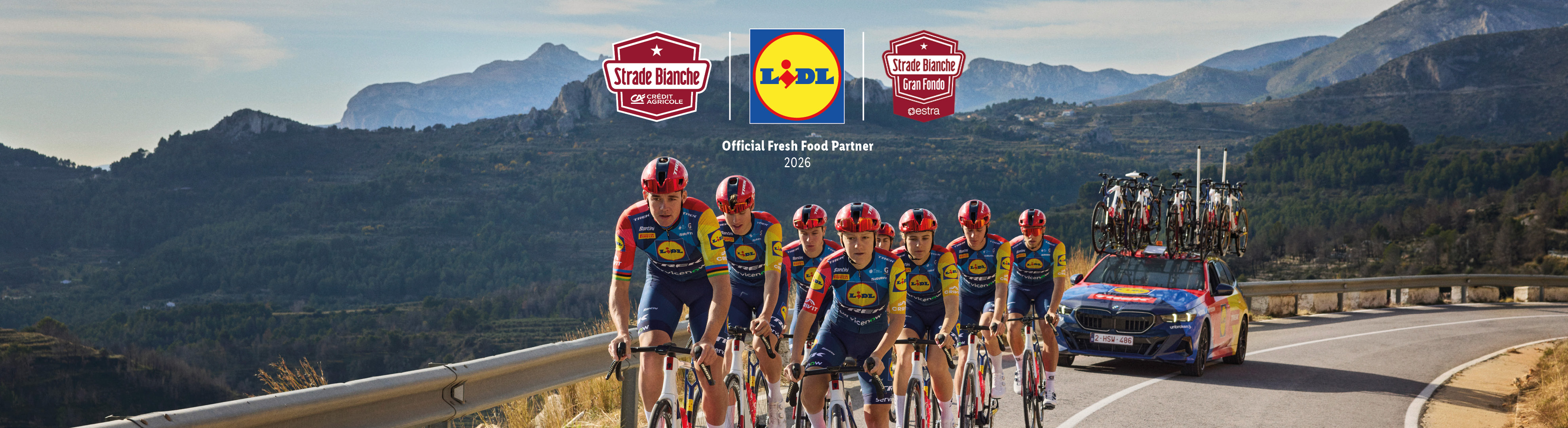 Ciclisti in maglia blu e gialla su una strada di montagna, con auto di supporto e loghi di sponsor.