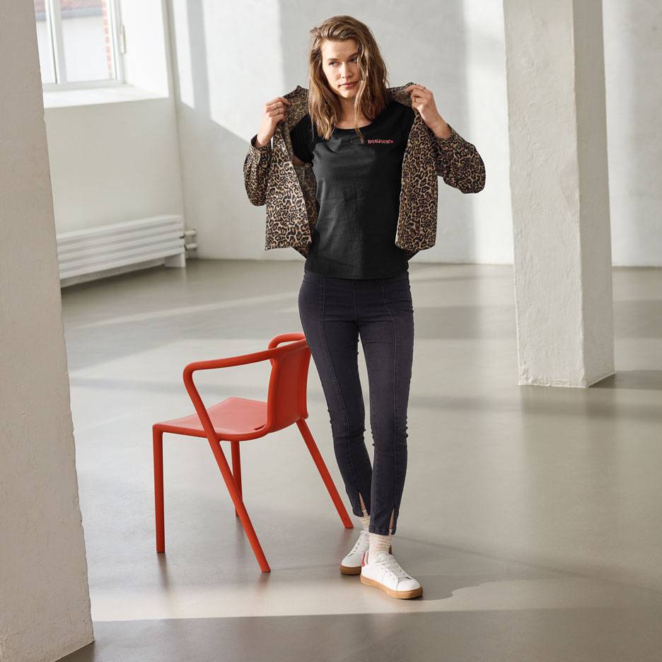 Donna in maglietta nera con scritta, jeans e giacca leopardata, in piedi accanto a una sedia rossa.