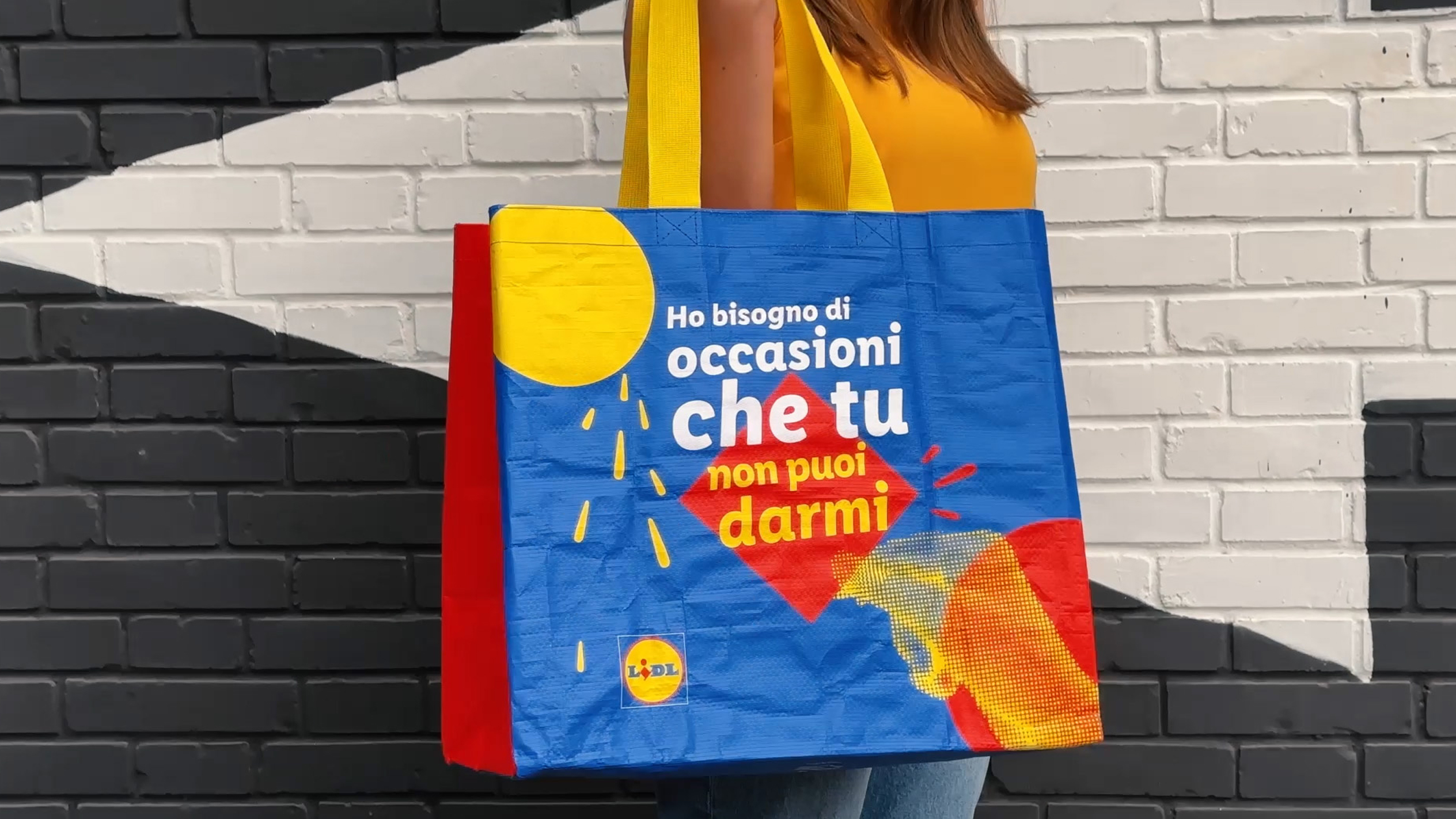 Una donna tiene una borsa della spesa Lidl con una grafica colorata e il testo 'Ho bisogno di occasioni che tu non puoi darmi'.