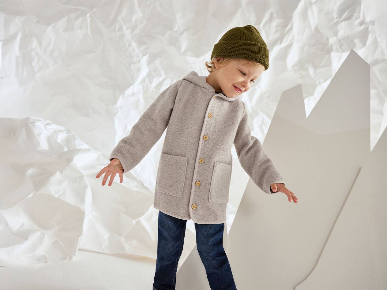 Bambino con giacca di lana grigia e berretto verde, sorridente su sfondo di carta stropicciata.