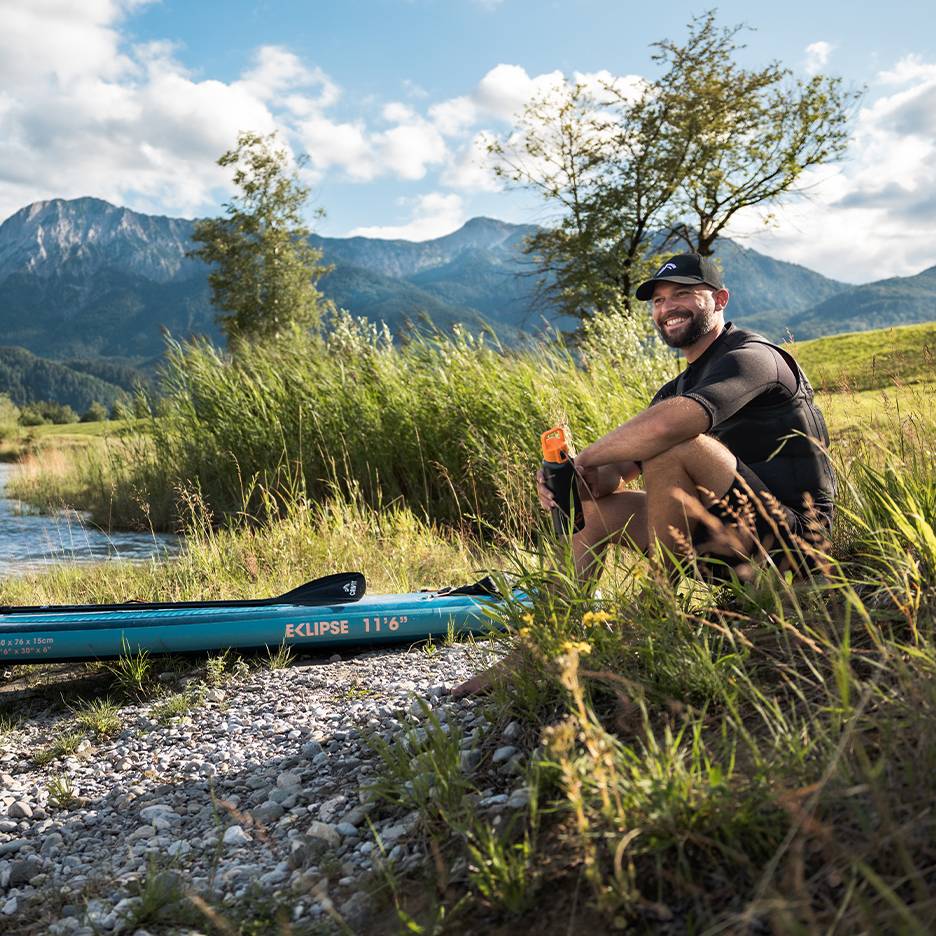 Uomo sorridente con cappellino e giubbotto di salvataggio, seduto accanto a una tavola da SUP blu in riva al fiume.