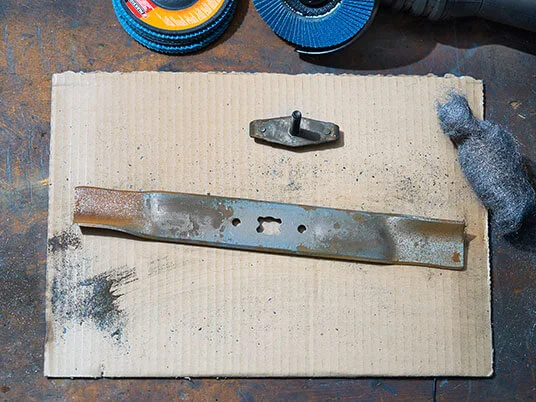 Lama di tosaerba arrugginita su cartone con dischi abrasivi e lana d'acciaio.