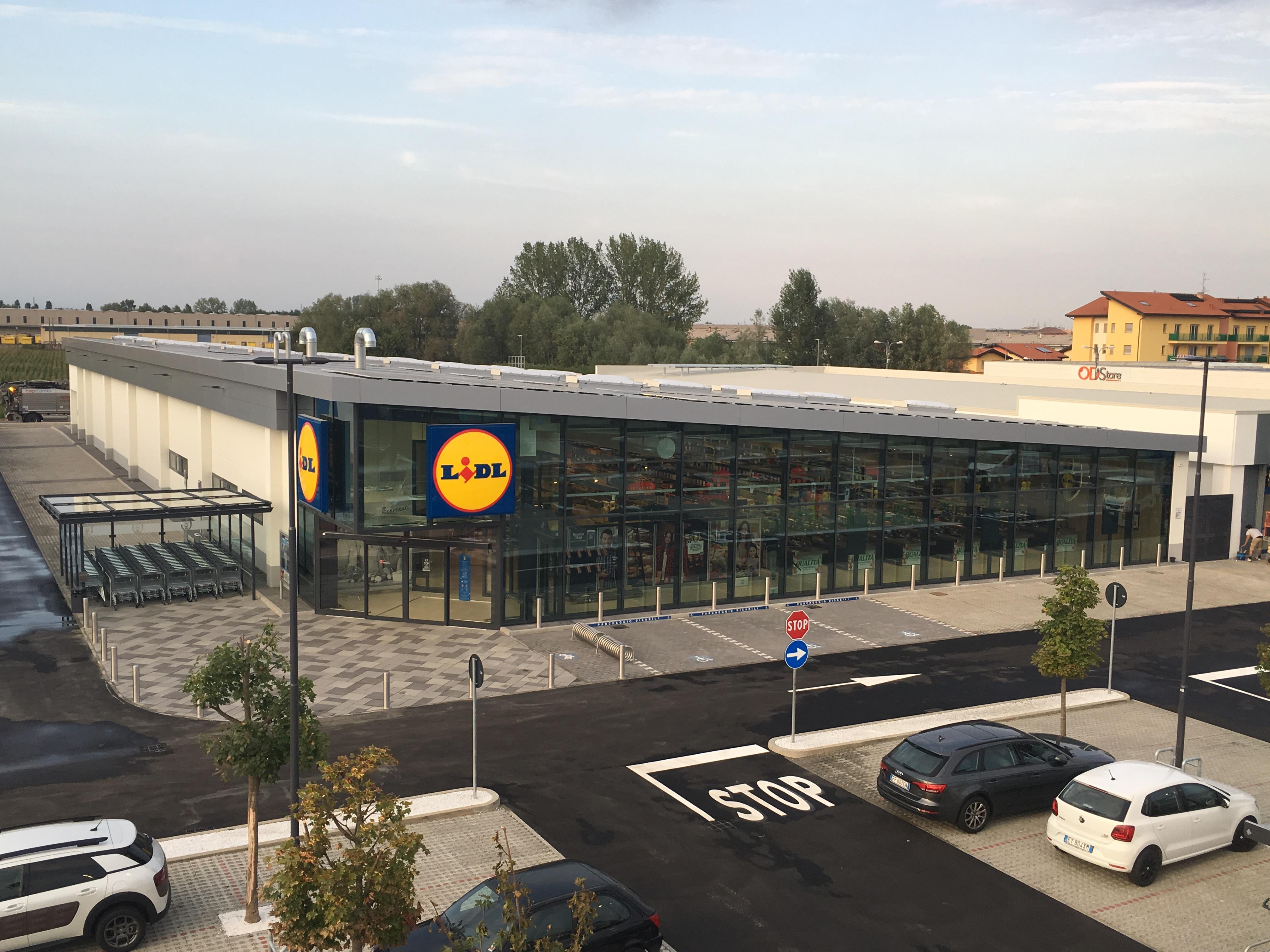 Esterno di un supermercato con parcheggio, carrelli e insegne luminose.