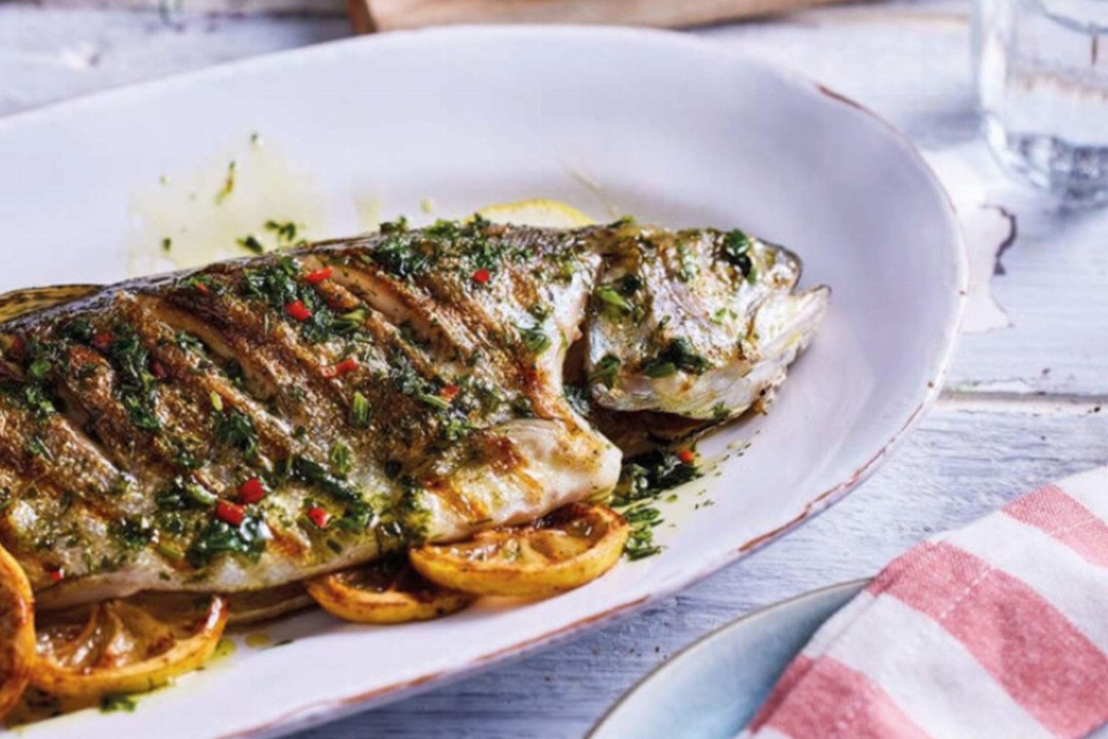 Pesce intero alla griglia con erbe e limone su un piatto bianco.