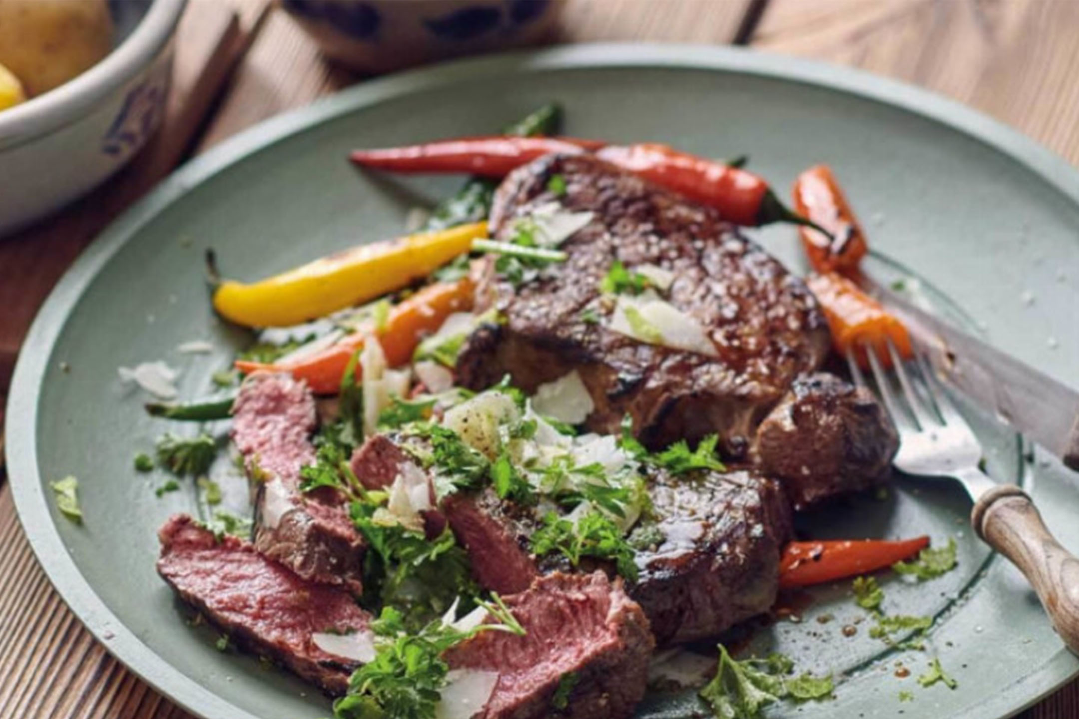 Bistecca alla griglia con peperoni, erbe fresche e scaglie di formaggio su un piatto verde.