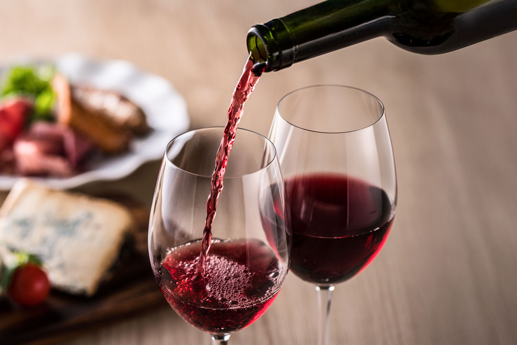 Vino rosso versato in un bicchiere, con antipasti sullo sfondo.