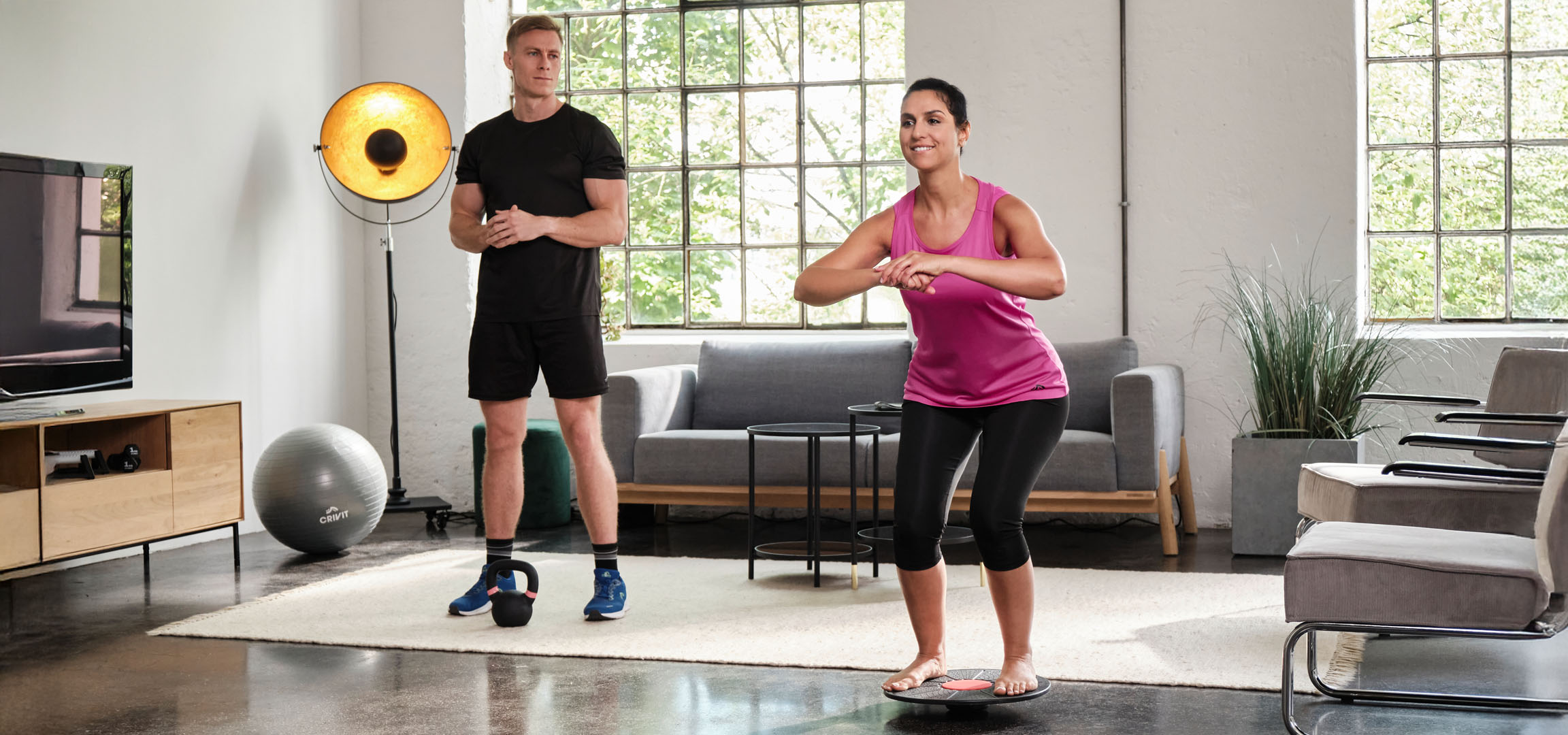 Donna e uomo che si allenano a casa con un balance board e un kettlebell.