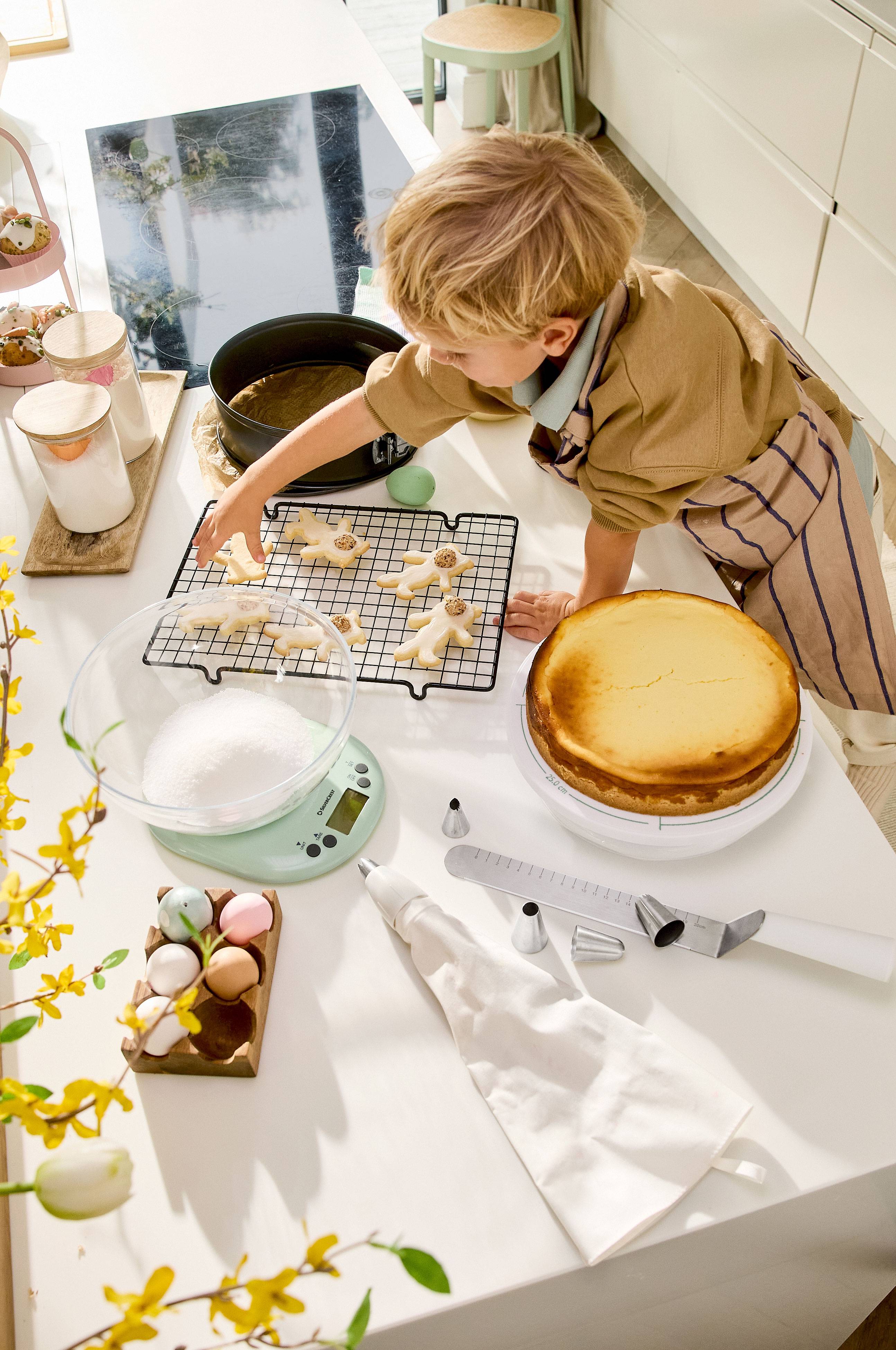 Un bambino decora biscotti di Pasqua, con una cheesecake e utensili da forno sul tavolo.