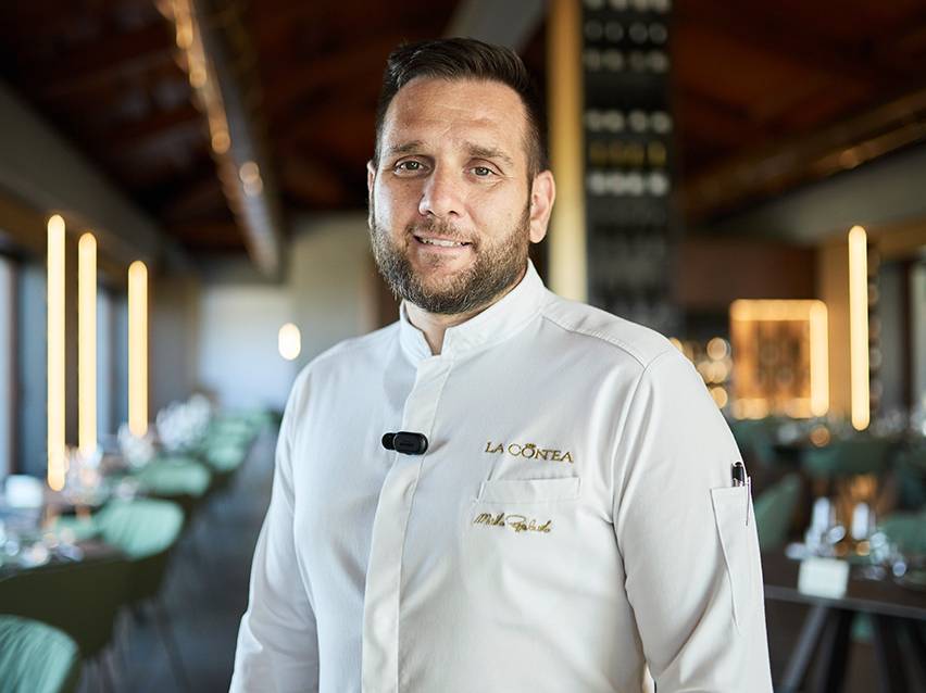 Chef sorridente in uniforme bianca con logo 'LA CONTEA' in un ristorante elegante.