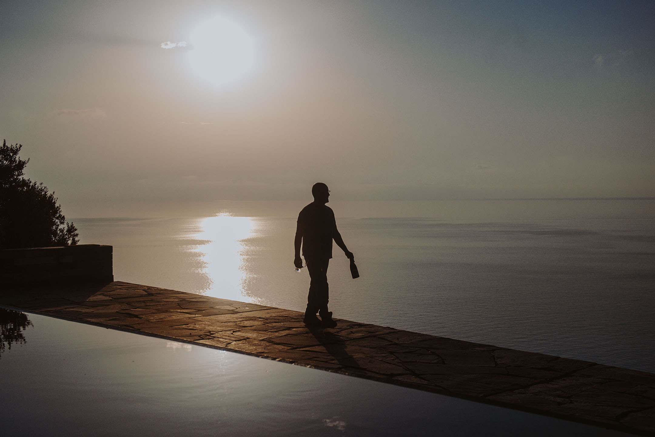 Silhouette di una persona con bottiglia e bicchiere a bordo piscina a sfioro al tramonto