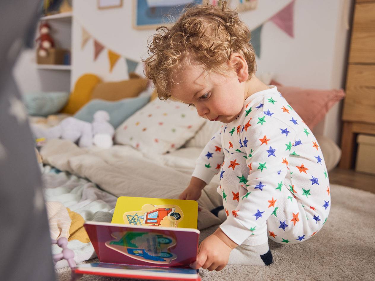 Un bambino piccolo in pigiama a stelle, seduto sul tappeto, legge un libro.