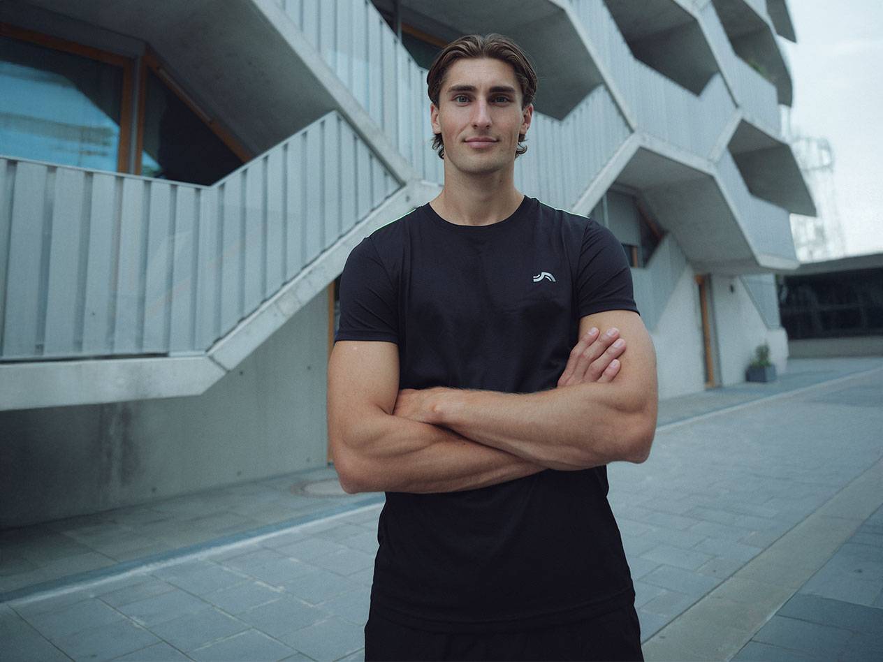 Uomo in maglietta nera con logo e pantaloncini neri, in piedi all'aperto con le braccia incrociate.