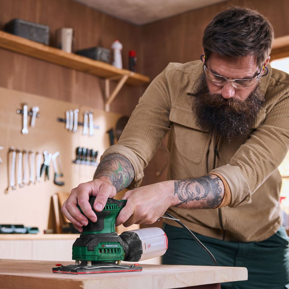 Uomo in officina leviga una tavola di legno con una levigatrice elettrica.