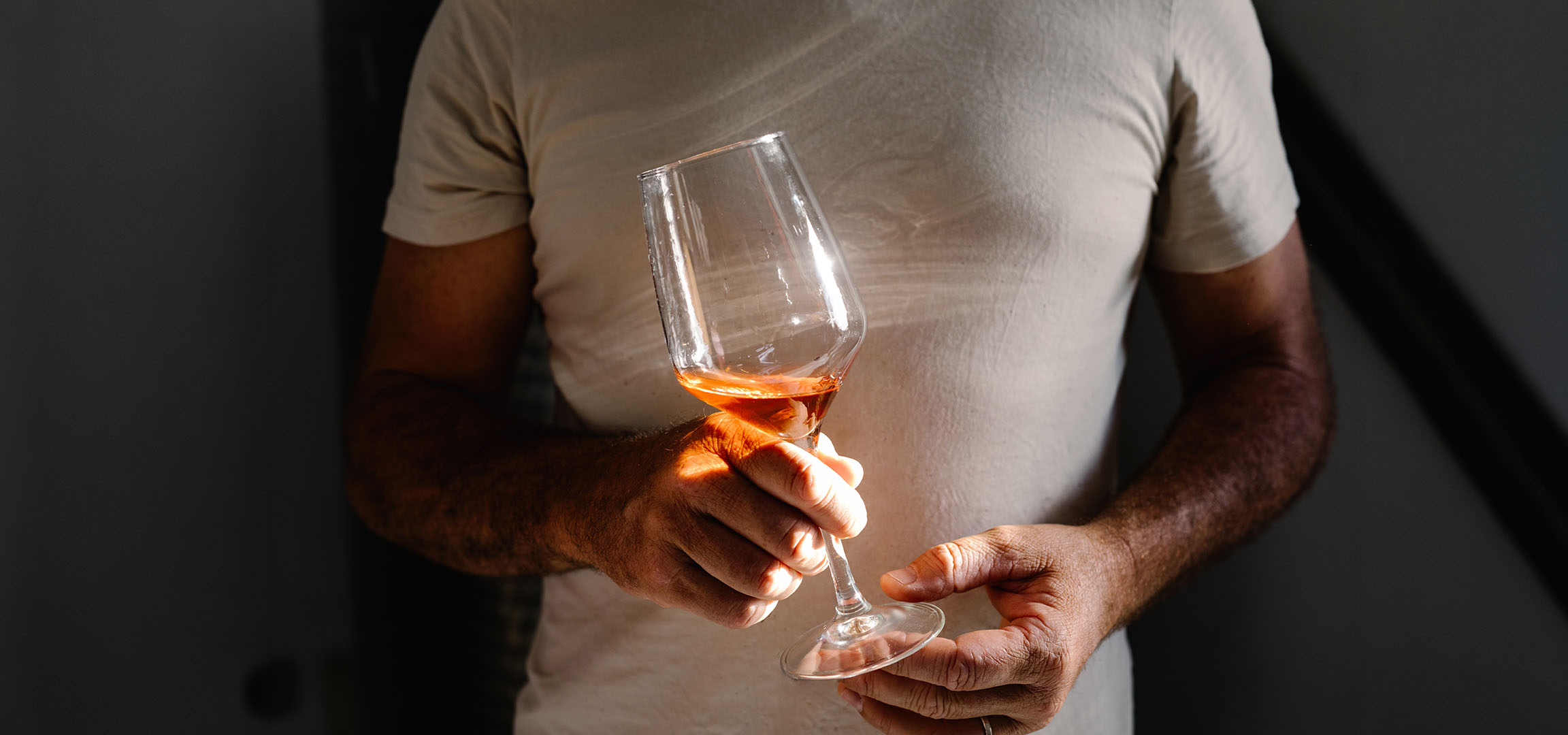 Uomo che tiene un bicchiere di vino rosato, illuminato dal sole, su sfondo scuro.
