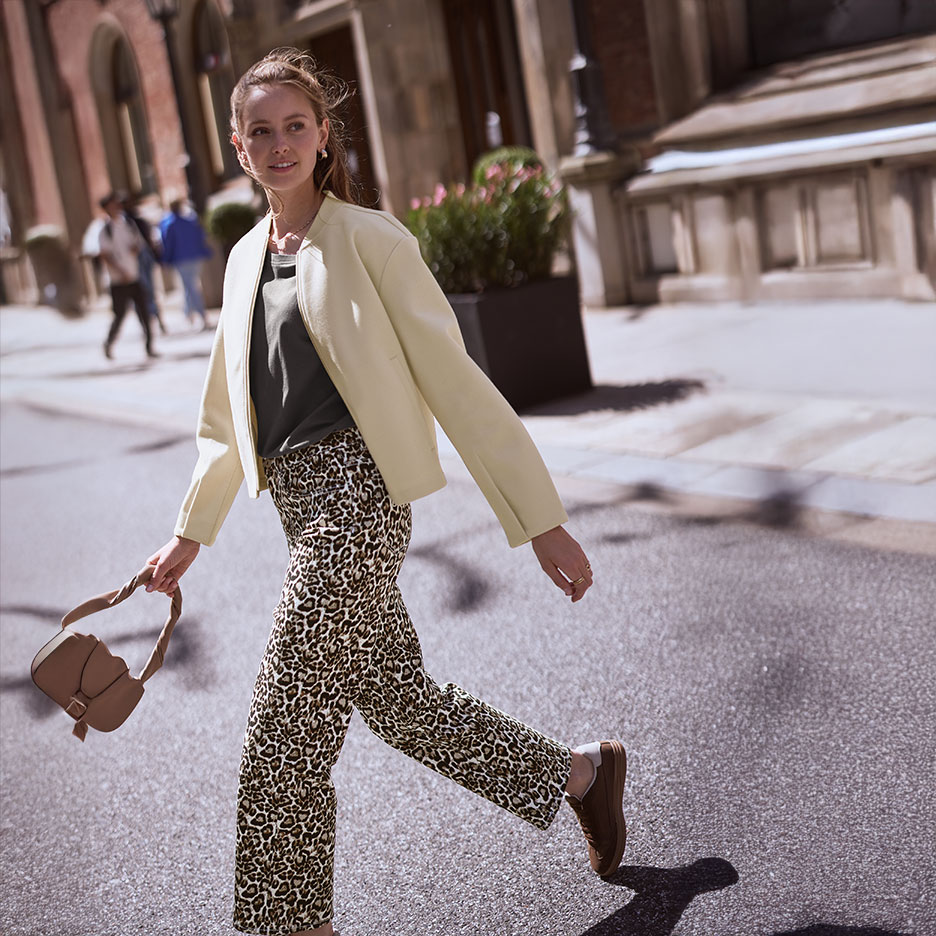 Donna in giacca beige, maglietta verde e pantaloni leopardati, cammina per strada con una borsa.