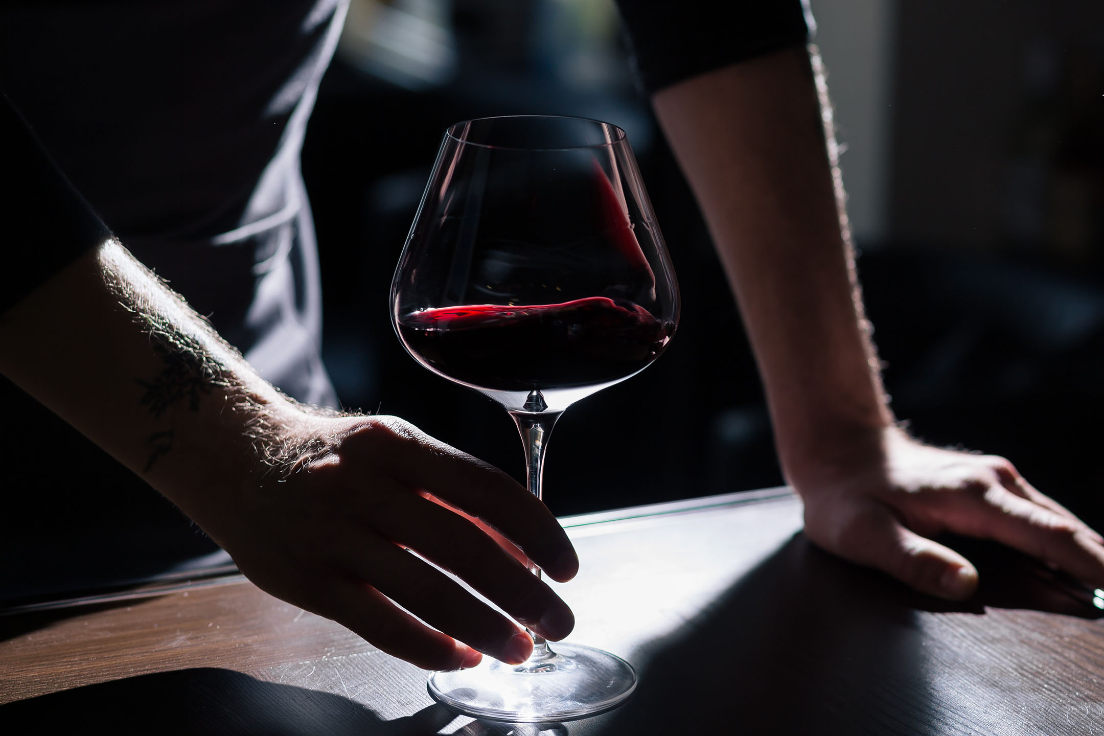 Mani tatuate di un uomo che tengono un bicchiere di vino rosso su un tavolo di legno.