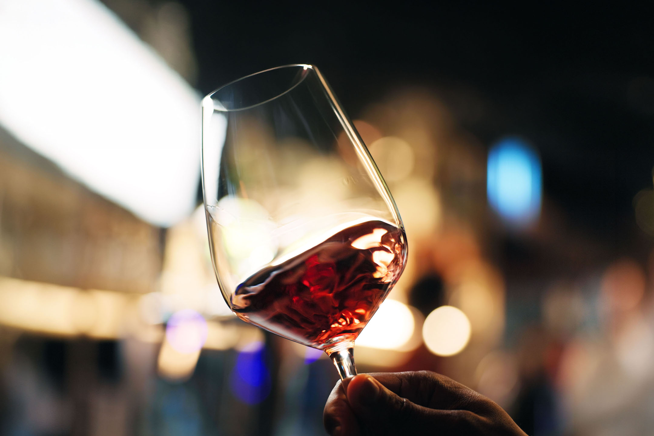 Bicchiere di vino rosso tenuto in mano, con luci sfocate sullo sfondo.