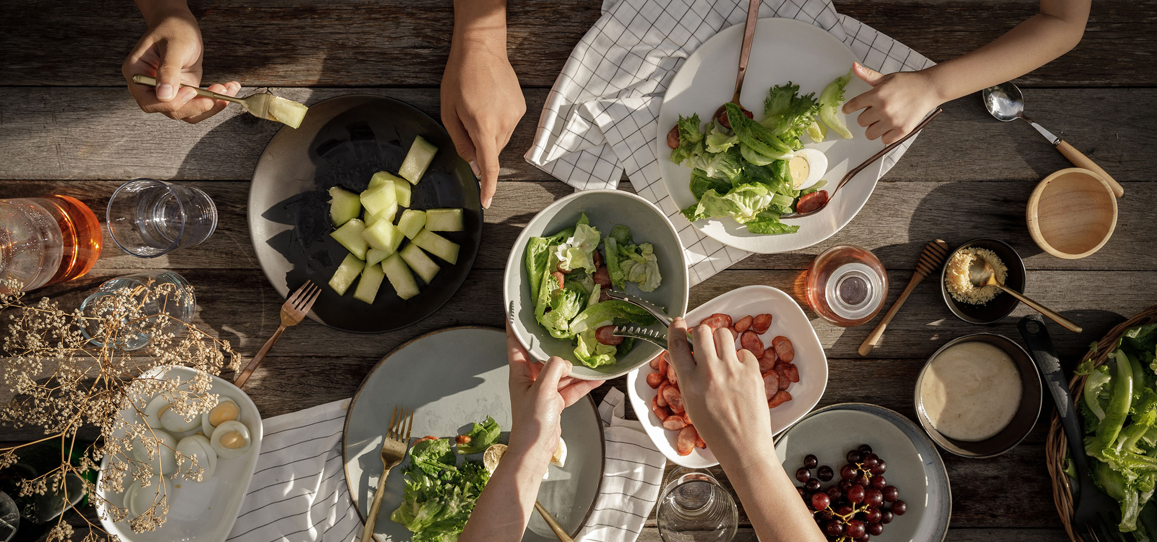 Vista dall'alto di un tavolo con insalata, salsicce e melone, persone che servono cibo.