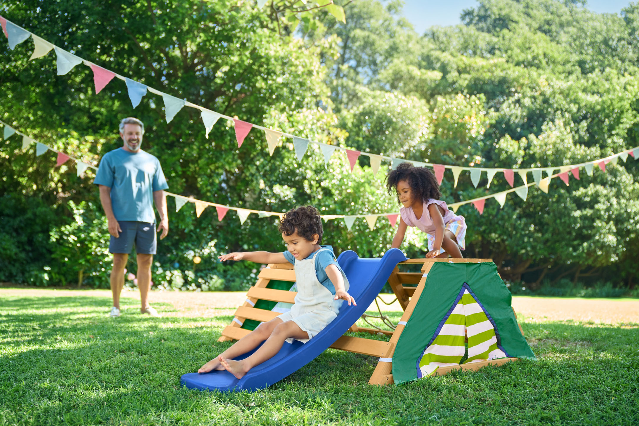 Bambini che giocano su un parco giochi in legno con scivolo in giardino, con il padre sullo sfondo.