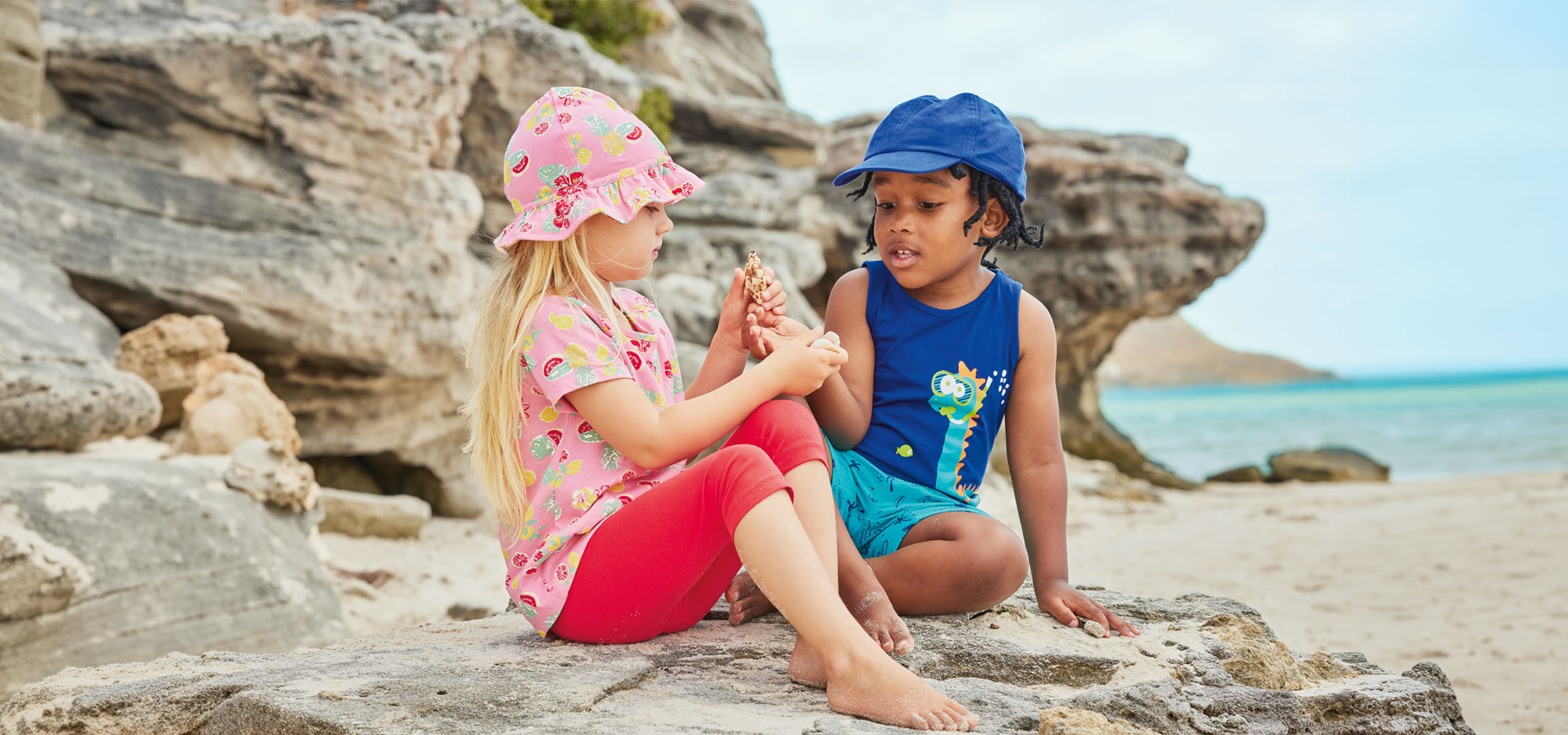 Due bambini in abiti estivi seduti su una roccia in spiaggia, con una conchiglia.