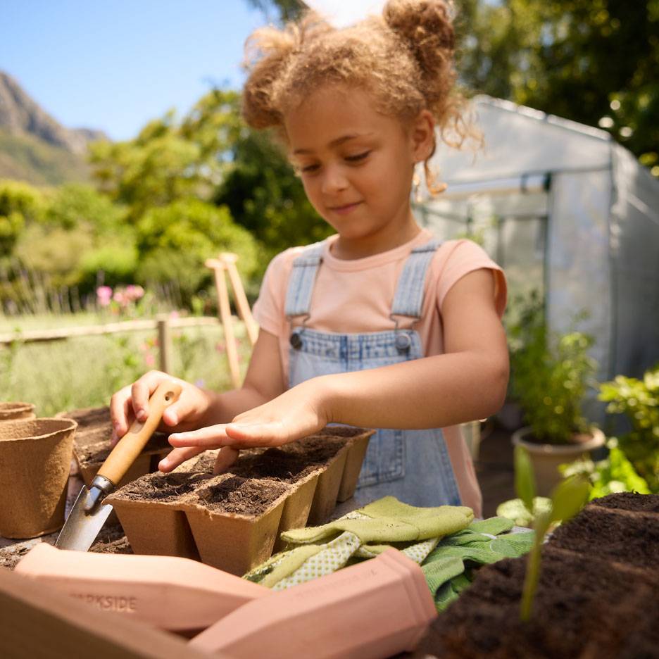 Bambina che pianta semi in vasetti di torba con attrezzi da giardinaggio e guanti.