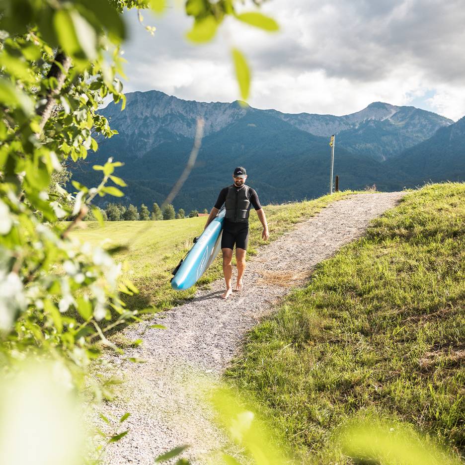 Uomo in muta e giubbotto di salvataggio che trasporta una tavola da paddleboard in un paesaggio montano.