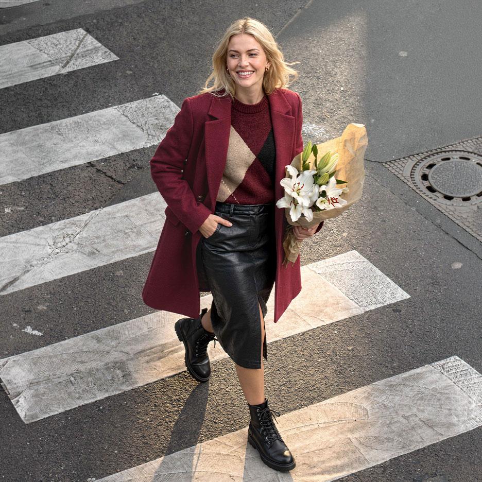 Donna sorridente con cappotto bordeaux, maglione a rombi e gonna in pelle, attraversa la strada con un mazzo di gigli.