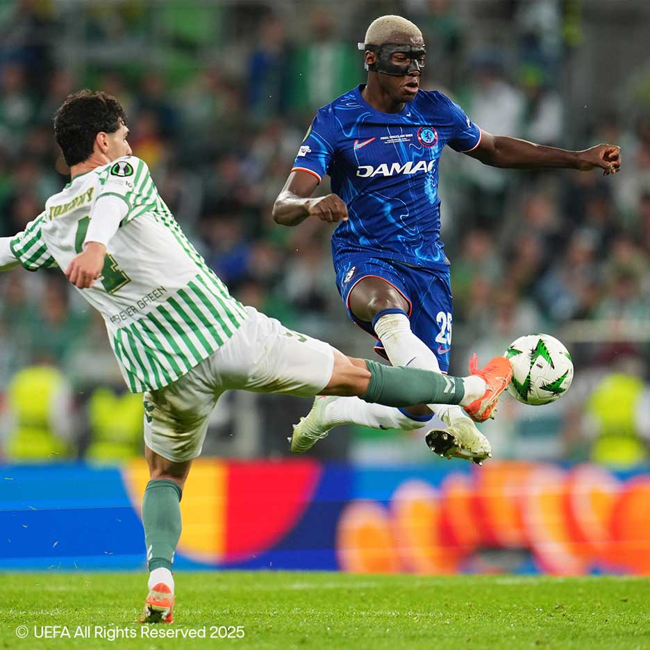 Due calciatori lottano per il pallone in campo, uno in bianco-verde, l'altro in blu con maschera.