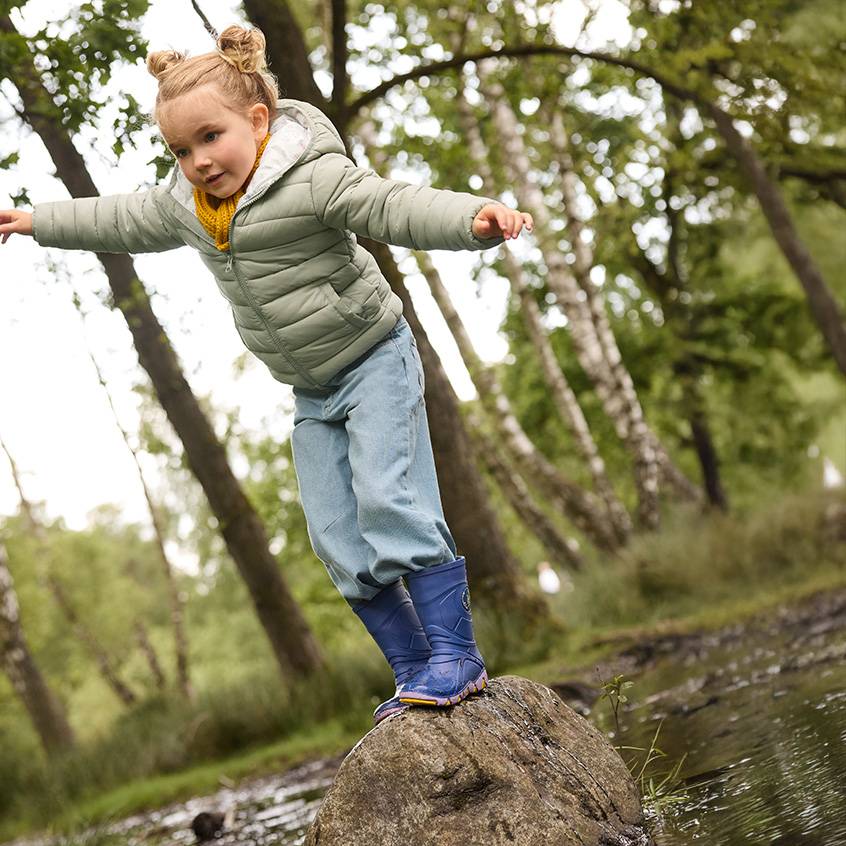 Bambina con giacca imbottita, jeans e stivali di gomma in equilibrio su una roccia nel bosco.