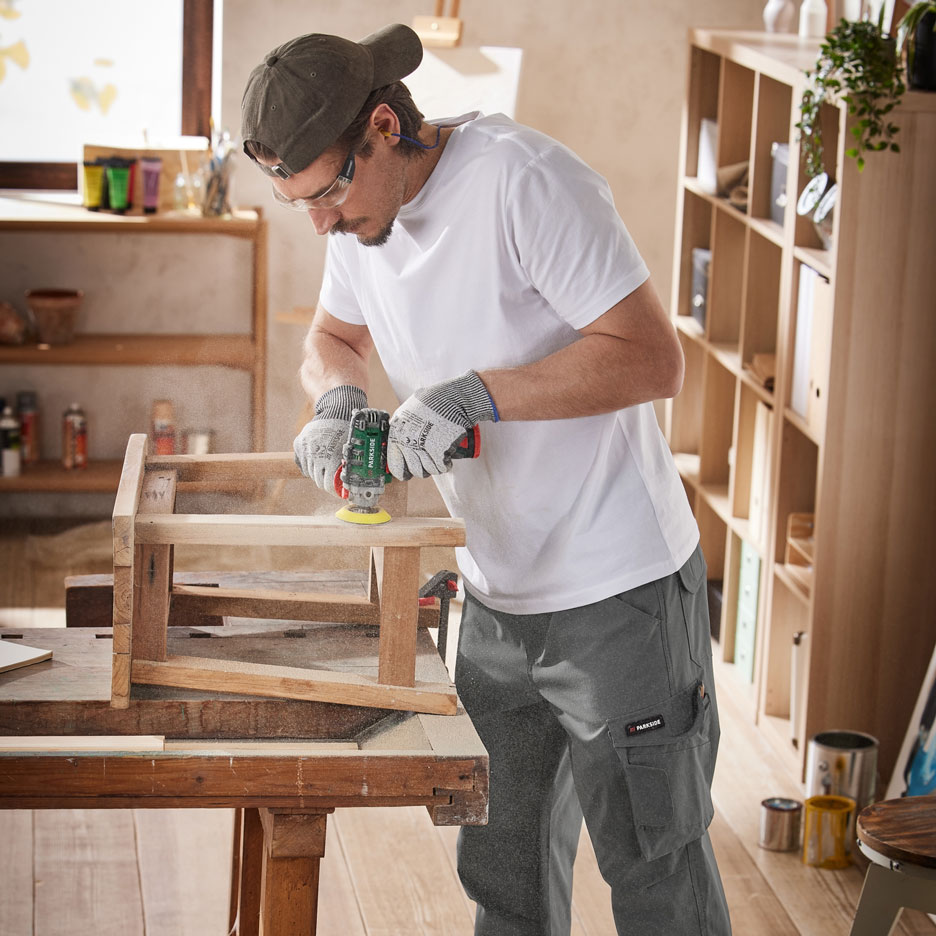 Uomo che leviga mobili in legno con un utensile elettrico, indossando pantaloni da lavoro e dispositivi di protezione.
