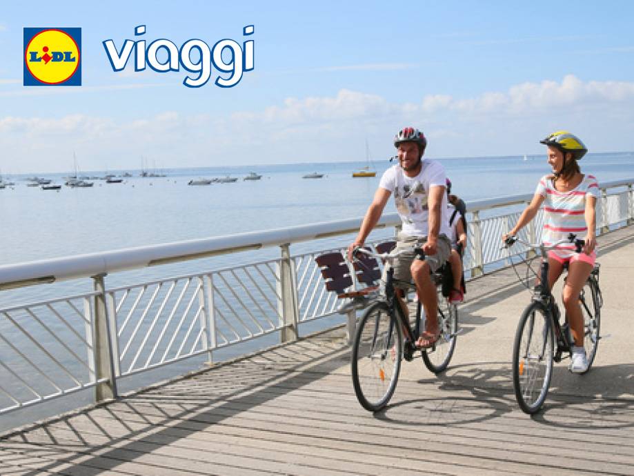 Coppia in bicicletta su un lungomare con barche, con logo 'viaggi'.