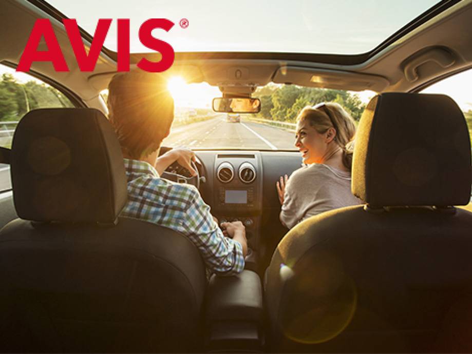 Coppia in auto su strada assolata, l'uomo guida e la donna si gira sorridendo.