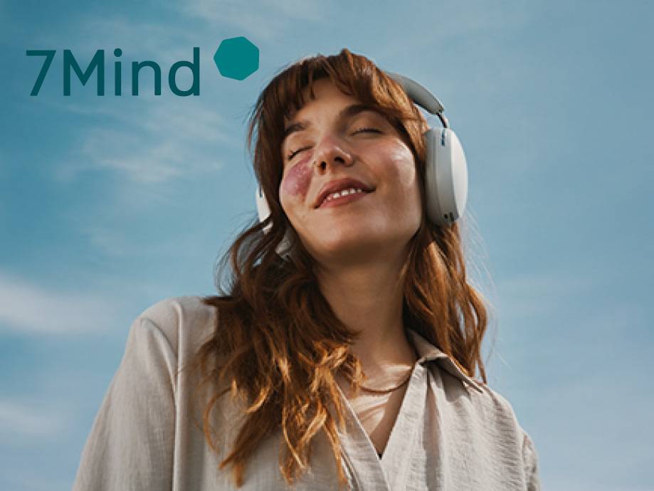 Una donna sorridente con cuffie bianche e il logo 7Mind su uno sfondo di cielo azzurro.