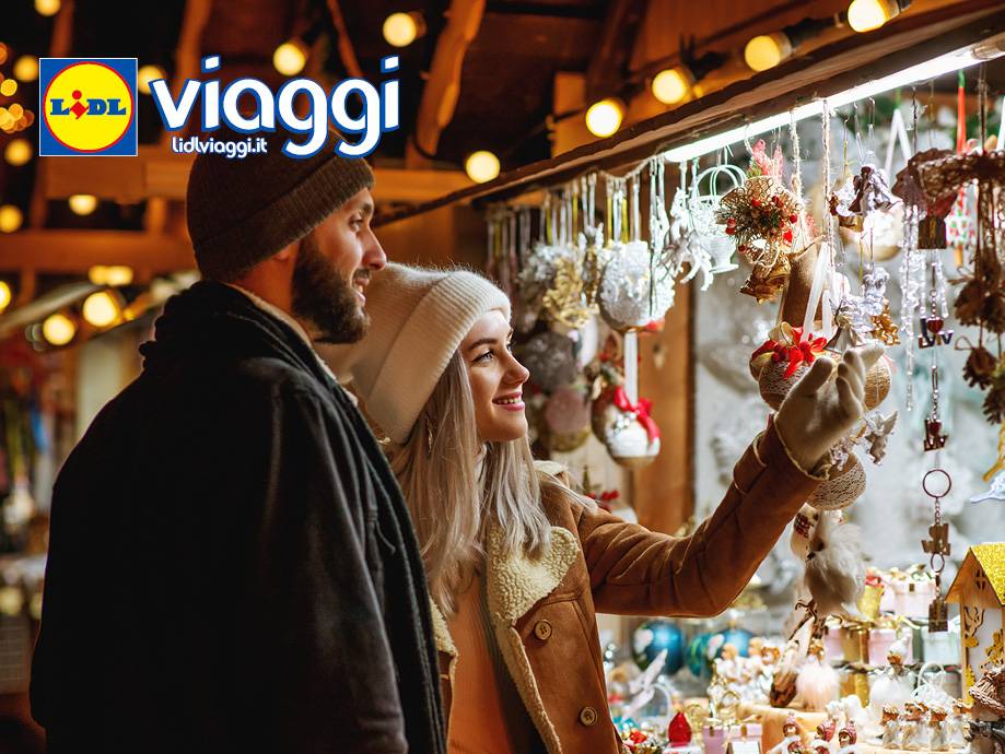 Una coppia sorridente guarda le decorazioni natalizie in un mercatino, con il logo Lidl Viaggi.