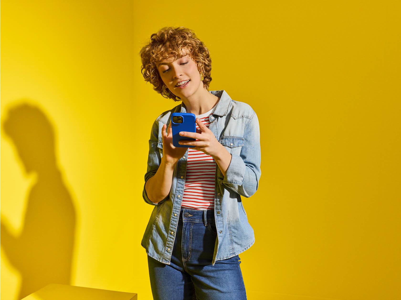 Una giovane donna con capelli ricci utilizza uno smartphone con custodia blu contro uno sfondo giallo.