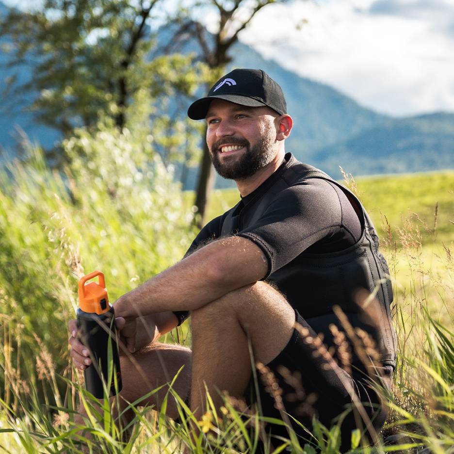 Uomo sorridente con cappellino nero e giubbotto in neoprene, tiene una borraccia arancione.