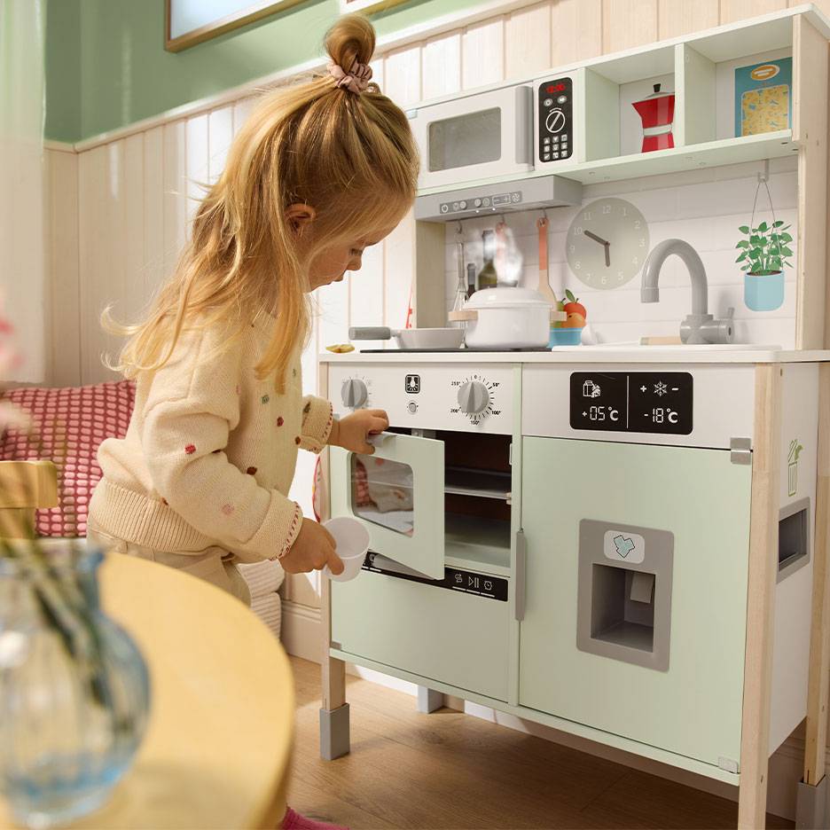 Bambina che gioca con una cucina giocattolo in legno, con forno, microonde e frigorifero.