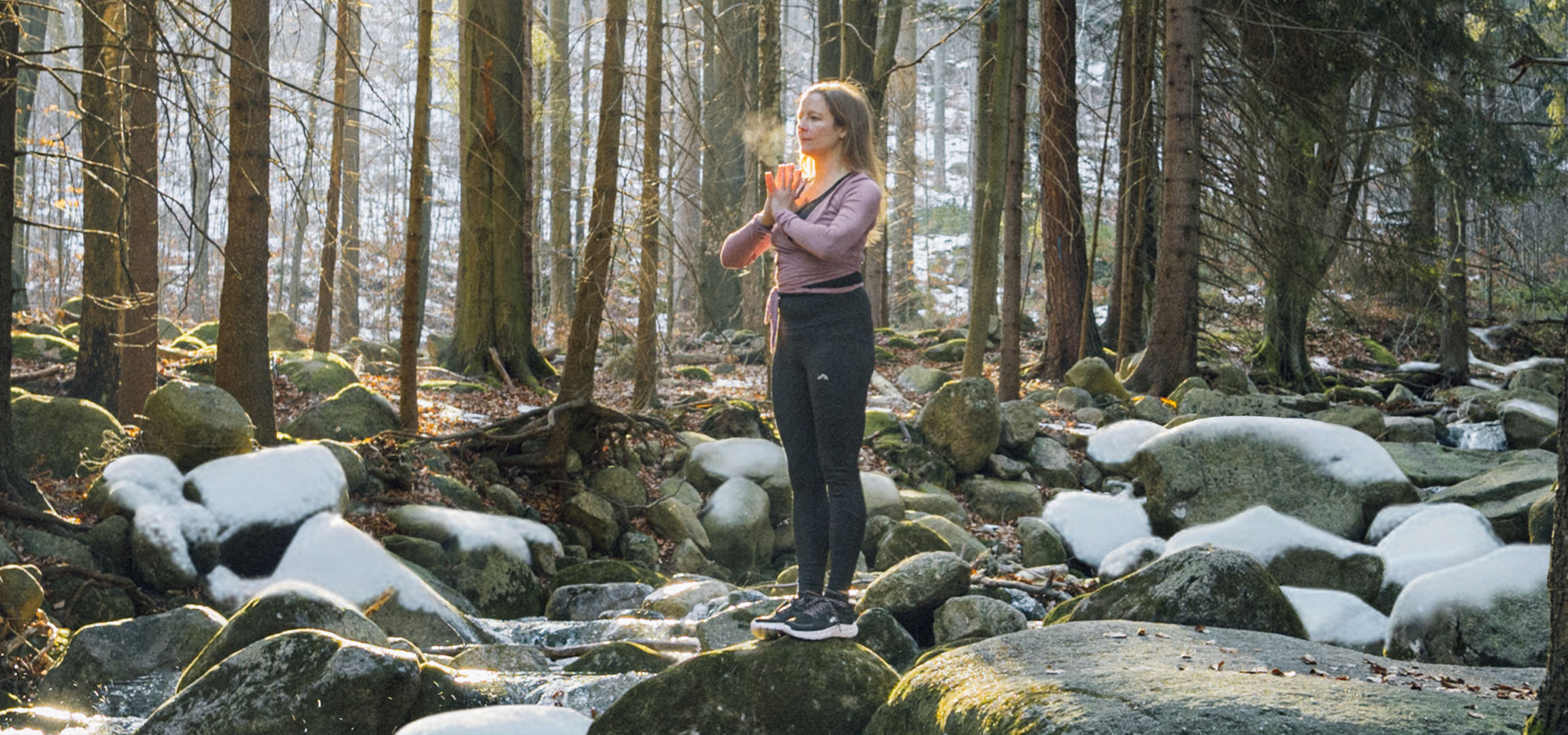 Donna in abbigliamento sportivo che fa yoga in una foresta innevata con un ruscello.