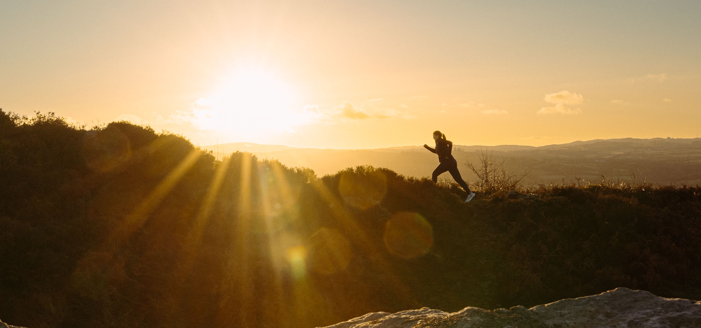 Donna che corre su una collina al tramonto, con raggi di sole e un paesaggio suggestivo.