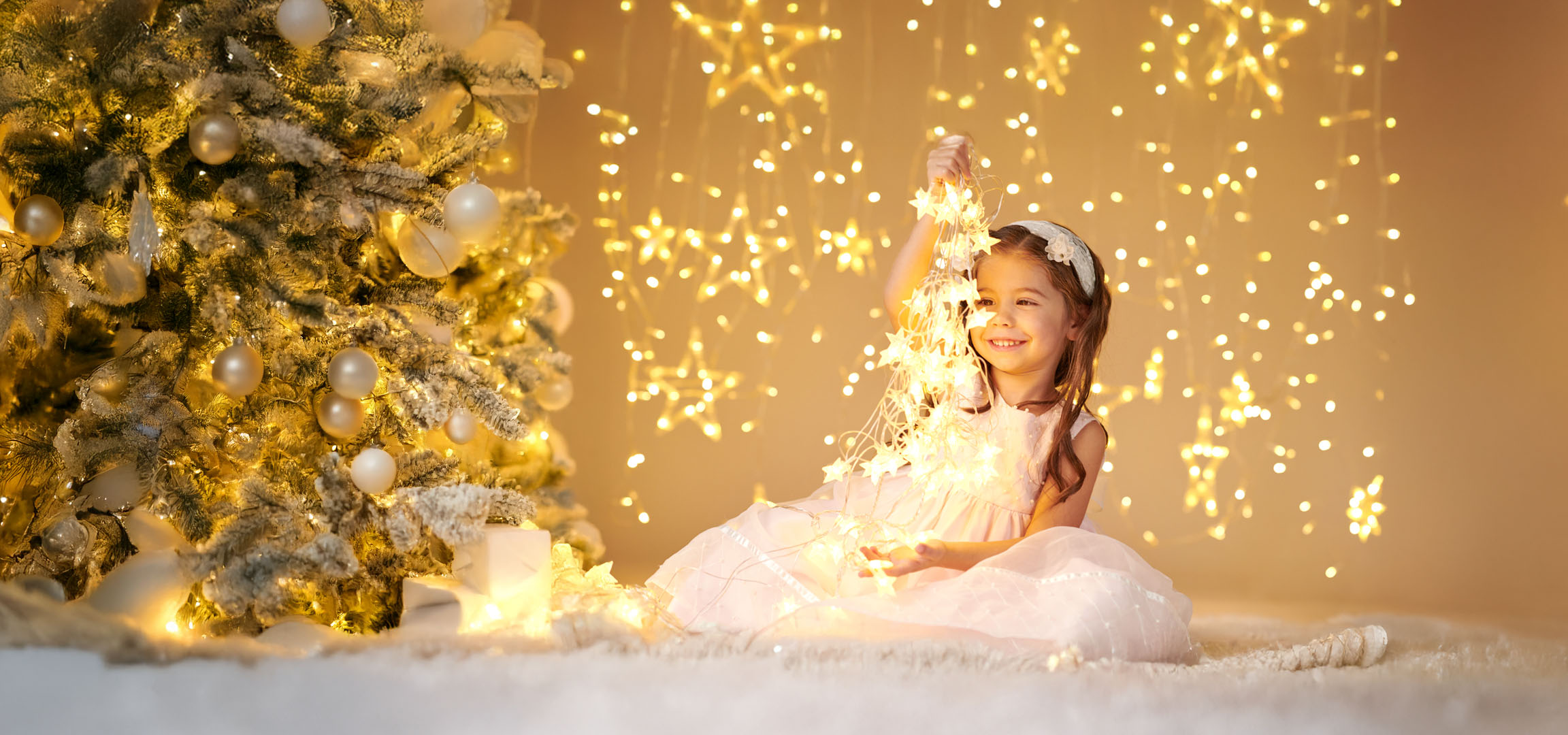 Bambina con luci di Natale davanti a un albero di Natale decorato.
