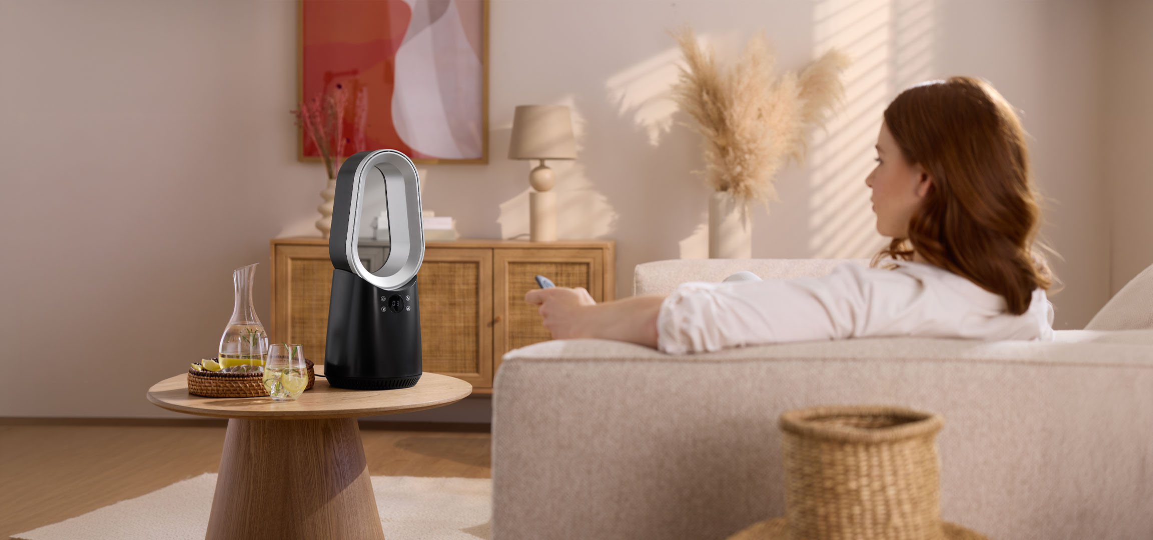 Donna sul divano con telecomando, ventilatore moderno e bevande sul tavolo.