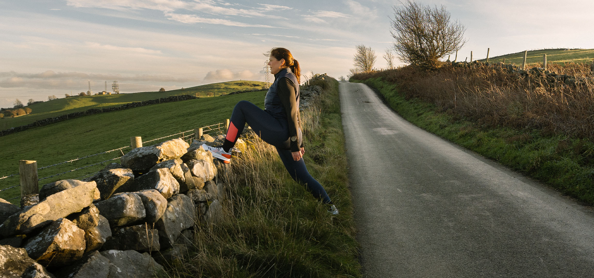 Donna che si allunga vicino a un muro di pietra su una strada di campagna al tramonto.