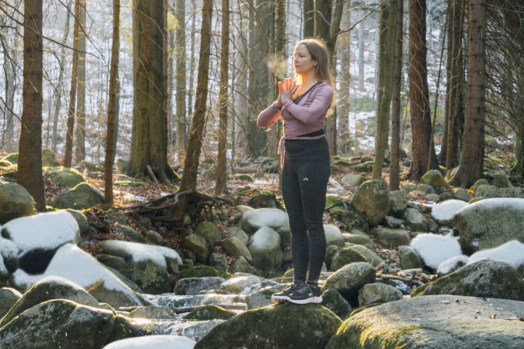 Donna in abbigliamento sportivo che pratica yoga in una foresta invernale vicino a un ruscello.