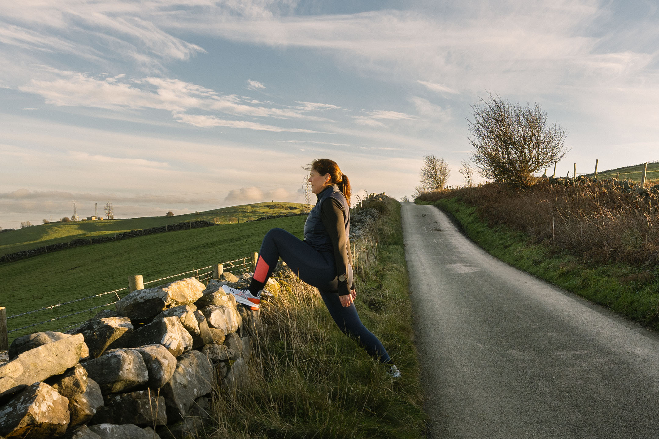 Donna in abbigliamento sportivo che si allunga su un muro di pietra lungo una strada rurale.