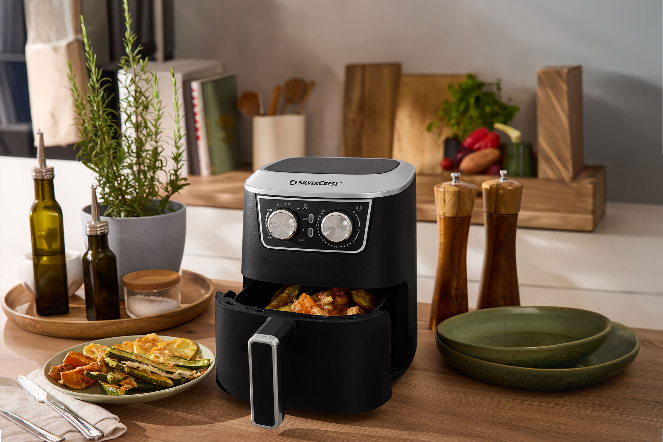 Friggitrice ad aria nera con verdure, spezie e piatti su un bancone di legno.